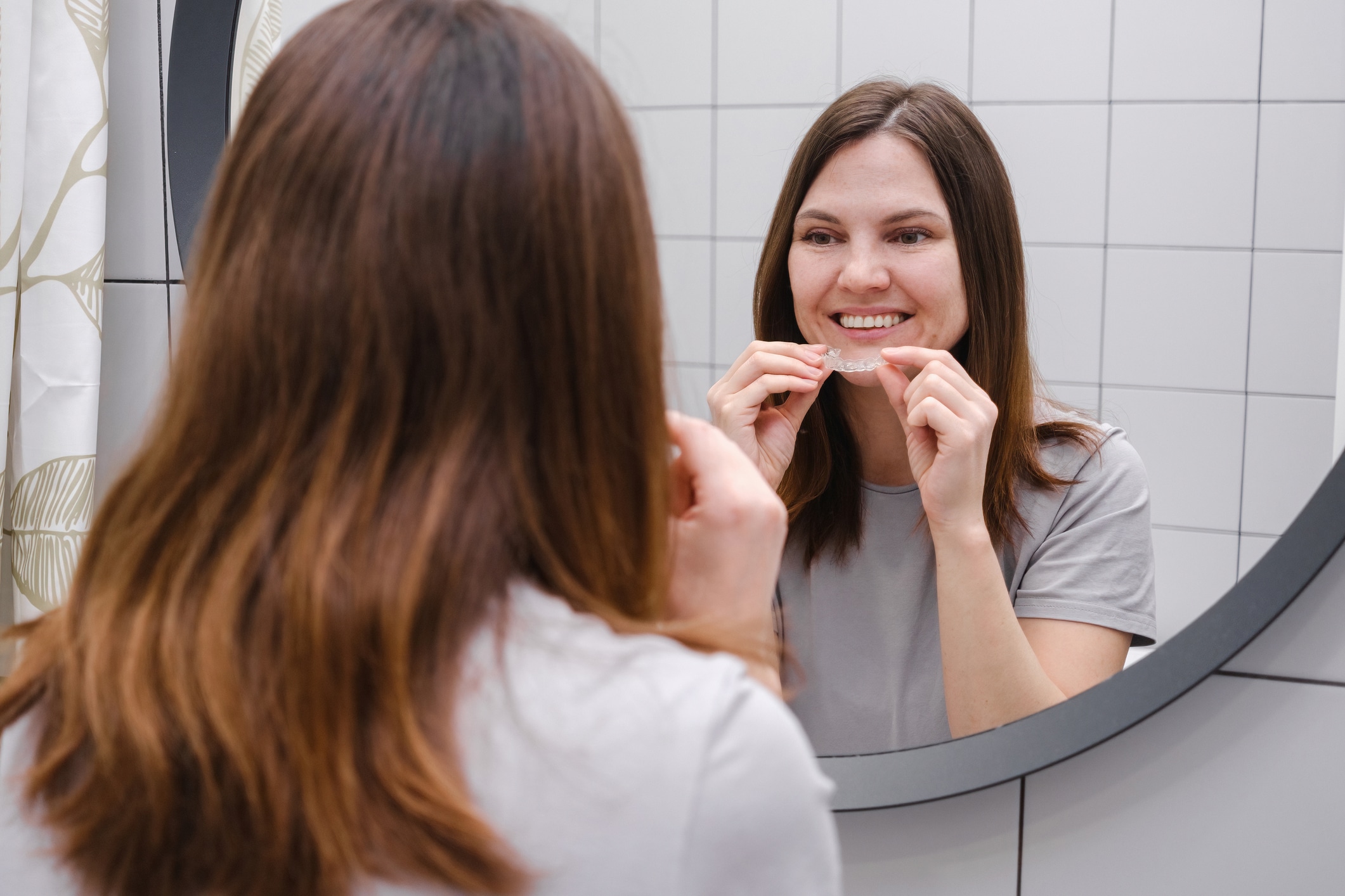 A widely smiling woman in front of a mirror in the bathroom holding invisible plastic teeth aligners in hands. Putting on braces. Beautiful and healthy smile.